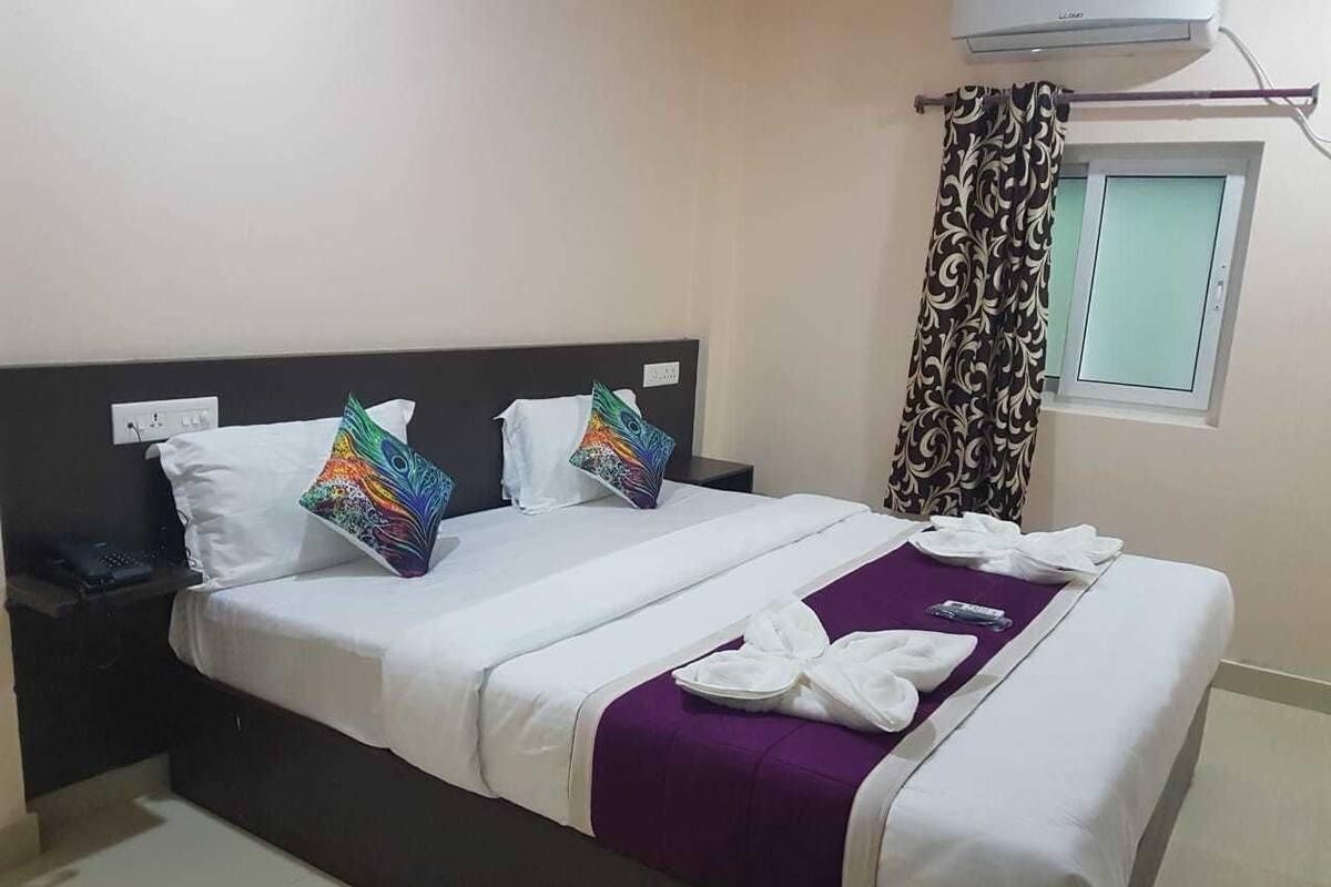 A neatly made bed with white bedding and a purple throw, a bedside table with a phone, and a window with curtains.