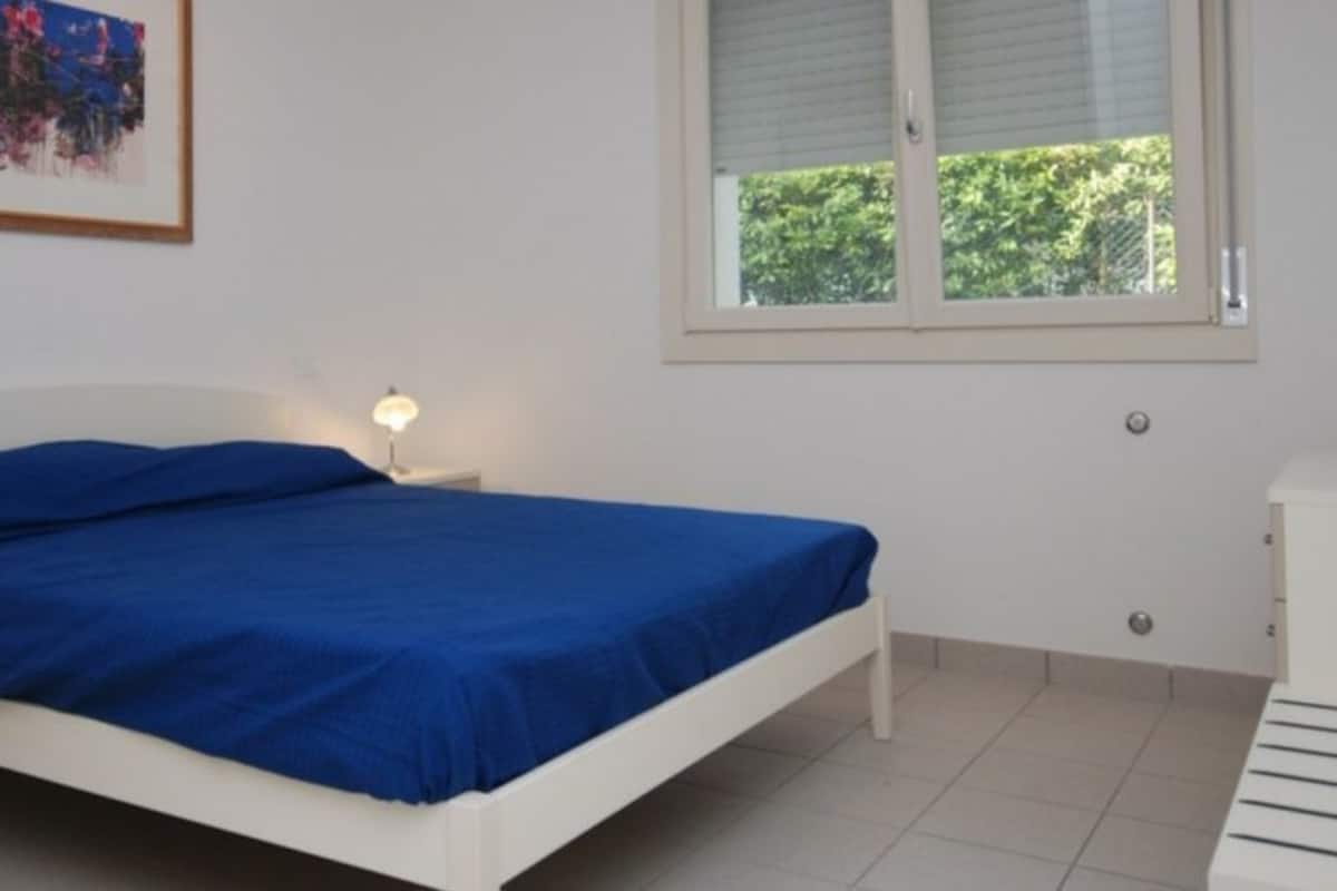 Quarto com uma cama de solteiro, mesa de cabeceira com abajur, janela com persianas e um quadro na parede.