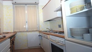 Cocina compacta con estufa a gas, lavamanos y refrigerador pequeño. Hay una ventana con cortinas y salpicadero de azulejos.