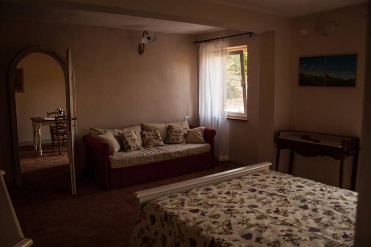 A cozy room with a bed, a sofa, and a table. There is a window with curtains, a painting on the wall, and a door leading to another room.