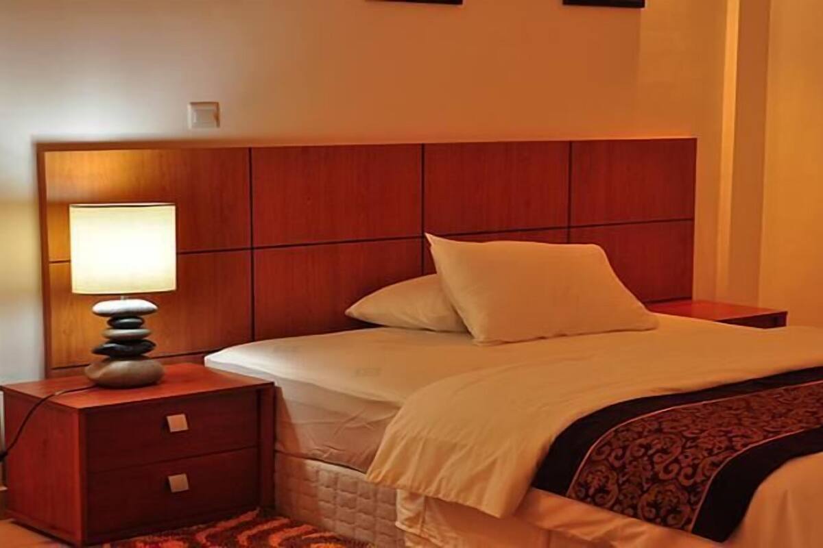 A hotel room with a wooden headboard, a bedside lamp, a bed with white bedding, and a patterned throw blanket.