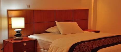 A hotel room with a wooden headboard, a bedside lamp, a bed with white bedding, and a patterned throw blanket.