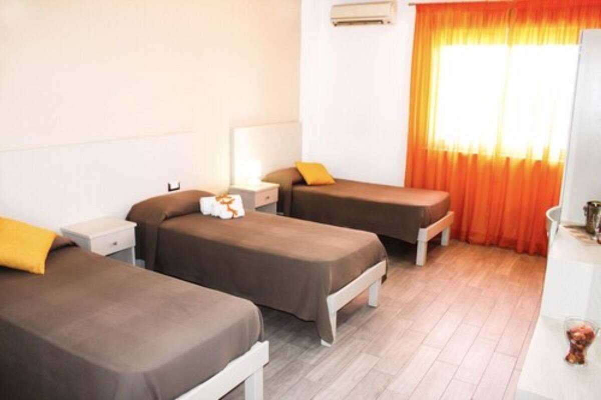 A hotel room with two single beds, a bedside table with a lamp, an air conditioning unit, and a window with orange curtains.