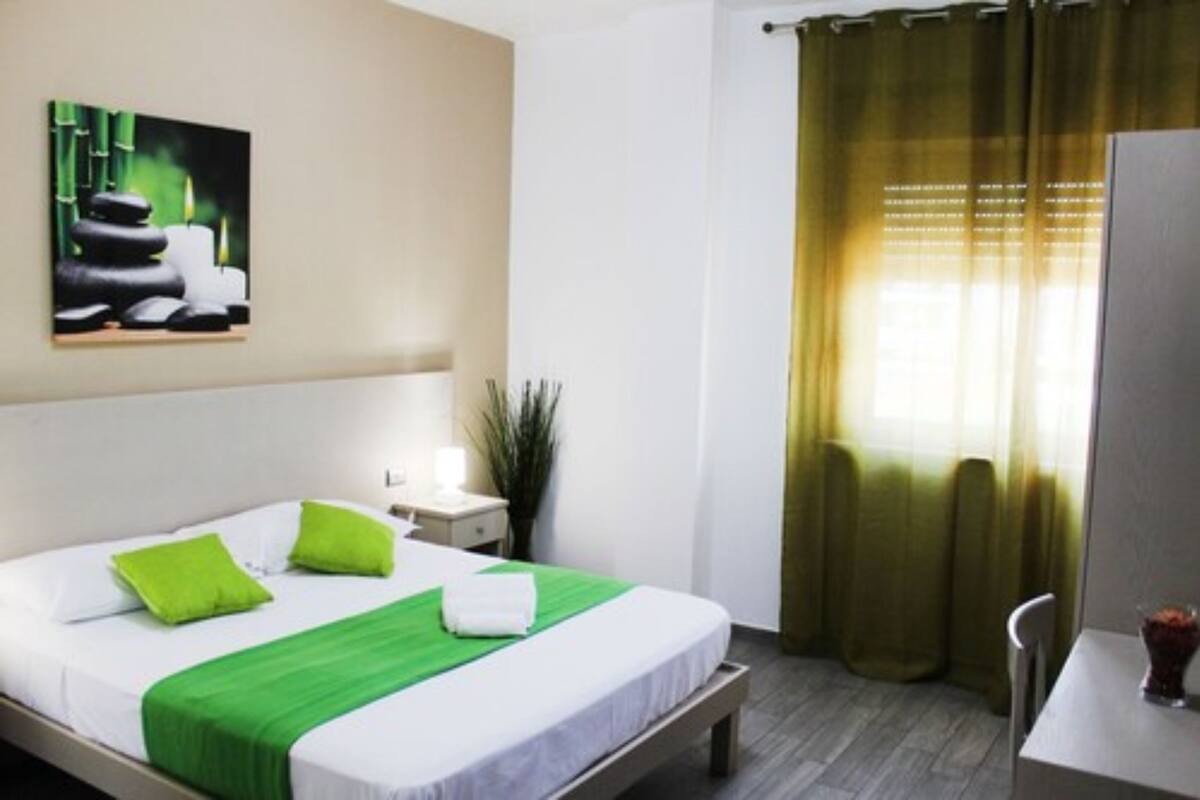 A hotel room with a bed, green pillows, a green bedspread, a painting on the wall, a nightstand, a lamp, a plant, a window with curtains, and a chair.