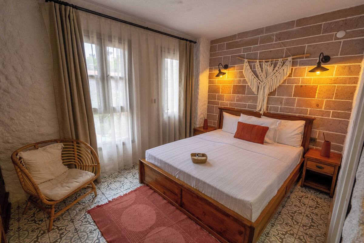 A bedroom with a wooden bed, a wicker chair, a patterned rug, and a brick wall with a macrame wall hanging.