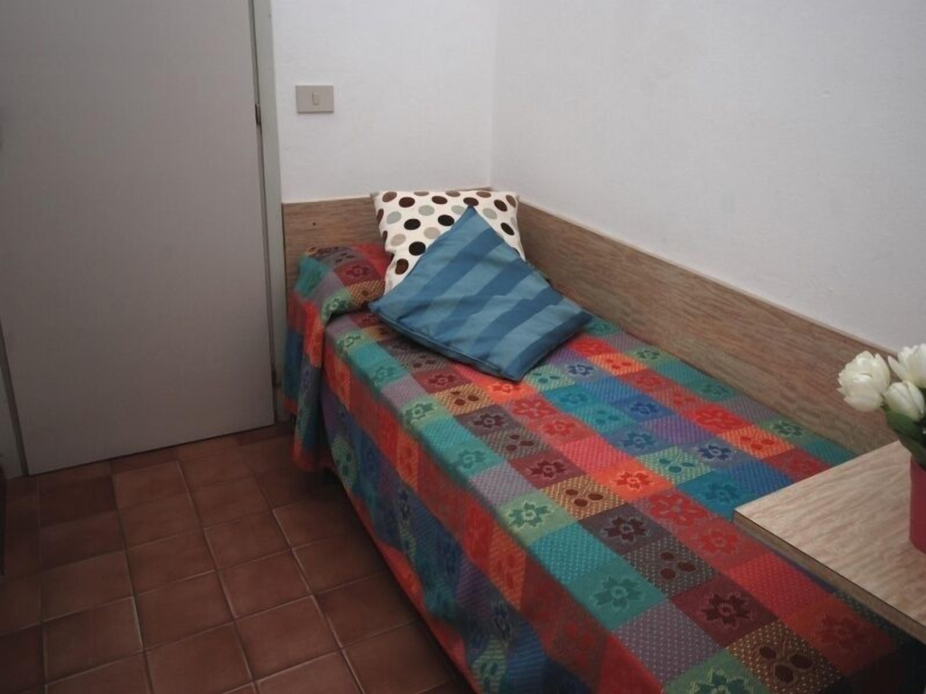 Une petite pièce, bien rangée, avec un lit, un oreiller, une petite table et un vase rempli de fleurs.