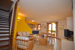A living room with a sofa, chairs, a dining table, and a TV. There is a staircase on the left and a kitchen in the background. - Villa Aurora (Lignano Sabbiadoro)