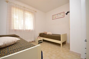 A bedroom with two single beds, a window with sheer curtains, a small bedside table with a book, and a wall-mounted lamp. - Villa Aurora (Lignano Sabbiadoro)