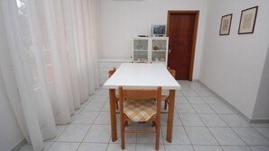A dining area with a white table and wooden chairs, a white cabinet with glass doors, and a door with two framed pictures on the wall. - Arcobaleno (Lignano Sabbiadoro)