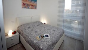 A bedroom with a bed, a nightstand, a lamp, a window with curtains, and a painting on the wall. - Arketipi (Lignano Sabbiadoro)