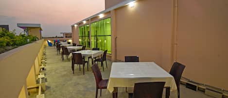 Un espacio para comer al aire libre con mesas y sillas, suelo de hormigón y un edificio con ventanas grandes al fondo.
