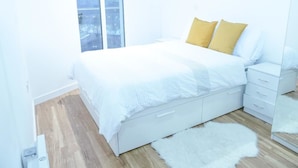 A bedroom with a bed, white nightstands, a white dresser, and a large window with a city view.