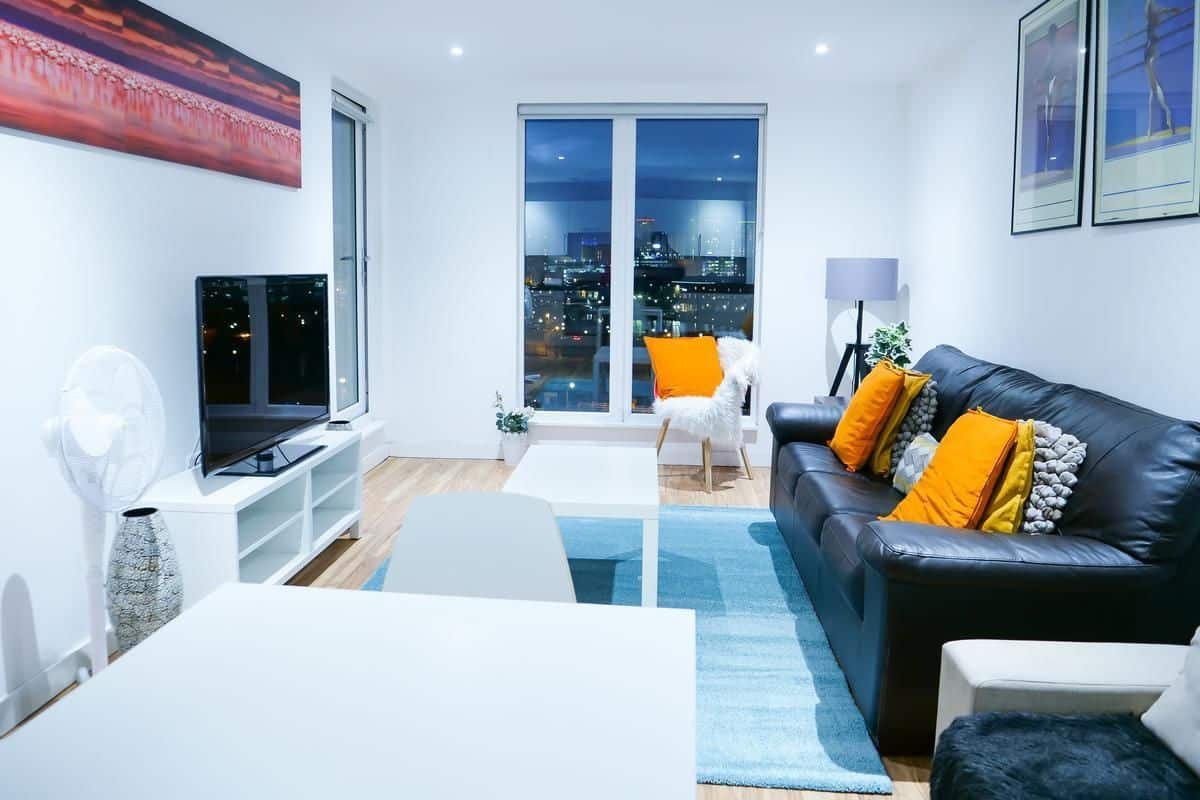 Una sala de estar moderna con vista a la ciudad, un televisor de pantalla plana, un sofá de cuero negro con cojines naranjas y una mesa de centro blanca.