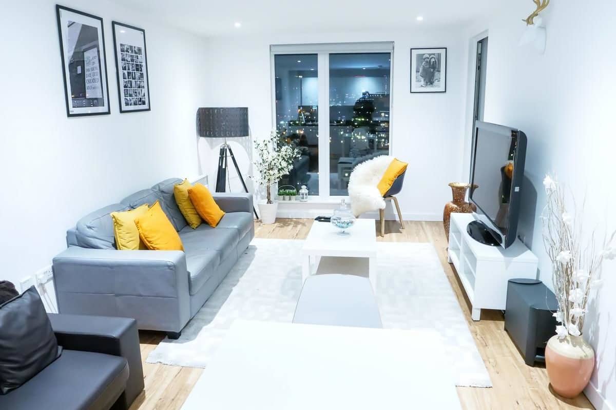 Un salón moderno con un sofá gris, cojines amarillos, una mesa de centro blanca, un sillón negro, un televisor y vista a la ciudad a través de la puerta de cristal.