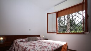 A bedroom with a bed, a nightstand, a window with a view of greenery, and a wooden headboard. - Villa Capinera (Lignano Sabbiadoro)