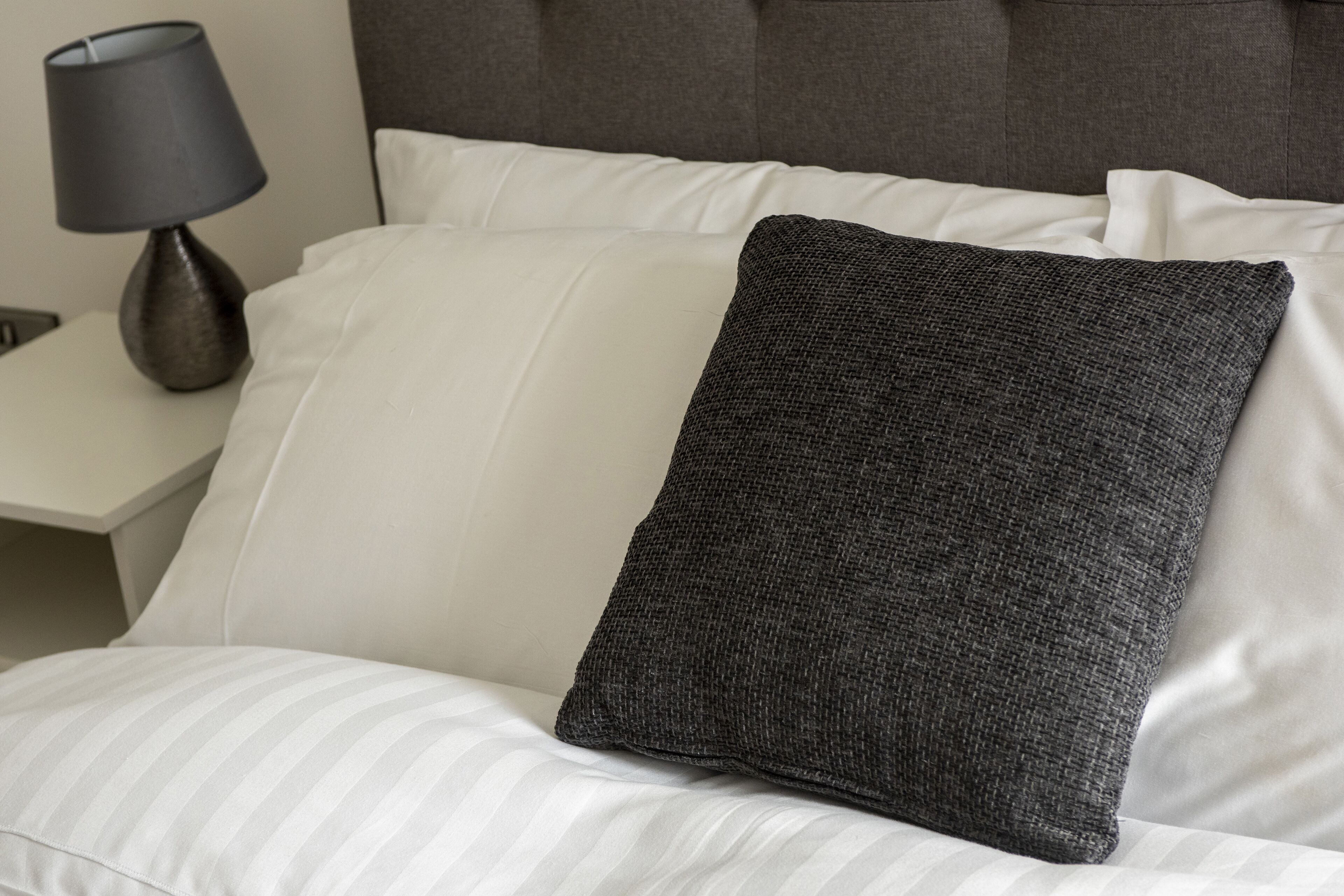 Ein Bett mit weiß gestreifter Bettwäsche, einem dunkelgrauen Kopfkissen und einer Lampe auf einem Nachttisch.