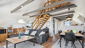 Un loft spacieux avec un plafond haut, des poutres apparentes, un canapé gris, une table à manger avec des chaises et un escalier menant à un niveau supérieur.
