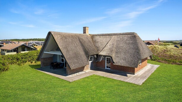 Une maison au toit de chaume, avec une base en briques, entourée d’une pelouse bien entretenue, située dans une zone rurale, avec d’autres maisons en arrière-plan.