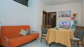 Een woonkamer met een oranje bank, houten stoelen, een eettafel met een gele tafelkleed, een televisie en een schilderij van zeedieren aan de muur.
