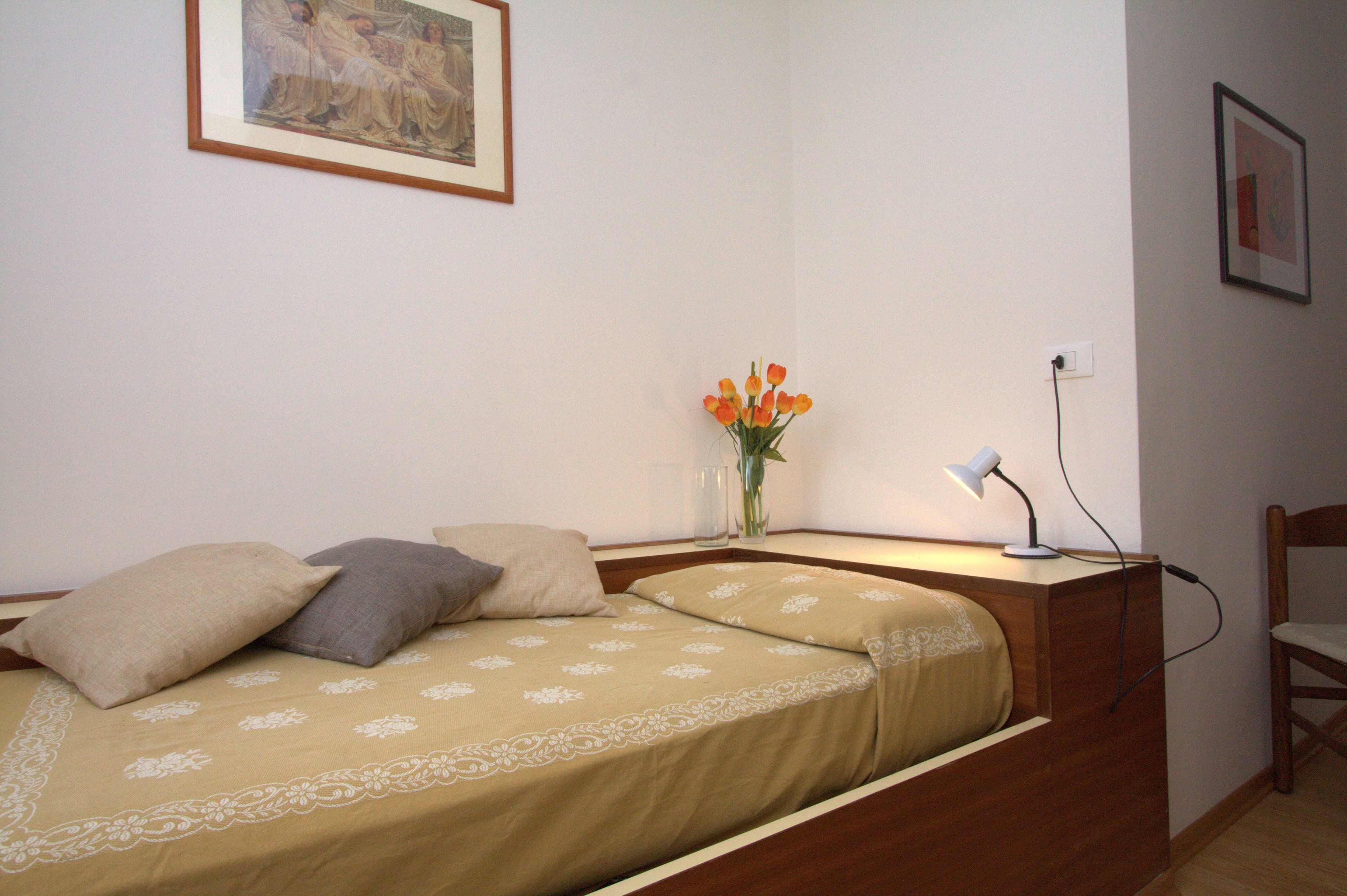 Una camera da letto con un letto, cuscini, un comodino con una lampada, un vaso con fiori e una sedia.