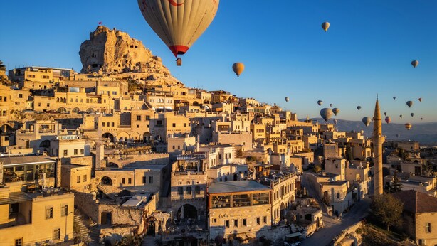 Paisagem urbana histórica com arquitetura tradicional, um minarete imponente e balões de ar quente no céu.