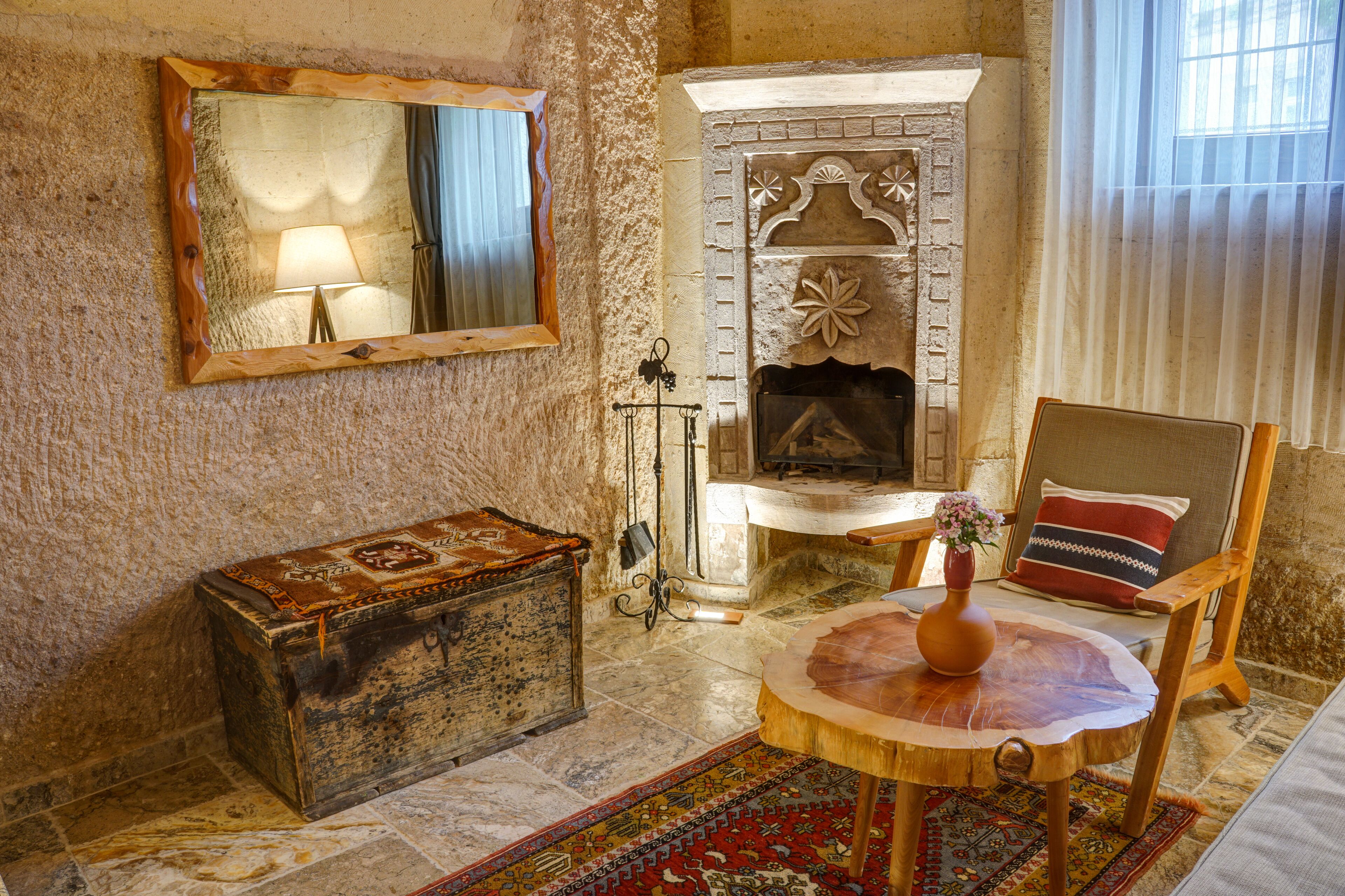 A rustic room with a stone fireplace, a wooden chest, a round wooden table with a vase, and a chair with a cushion.