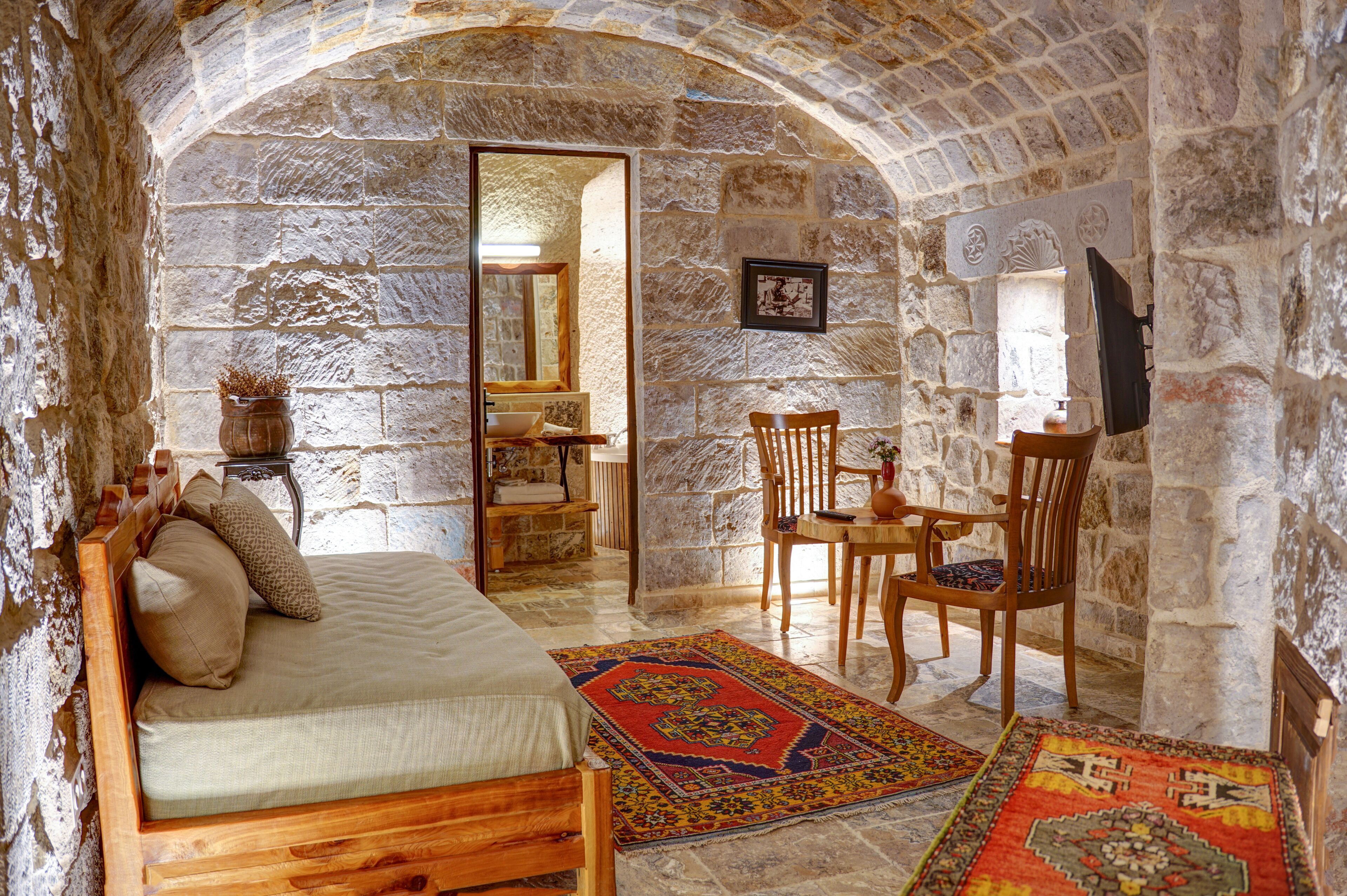 Une pièce aux murs de pierre, avec un lit, une chaise en bois, une table et un miroir.