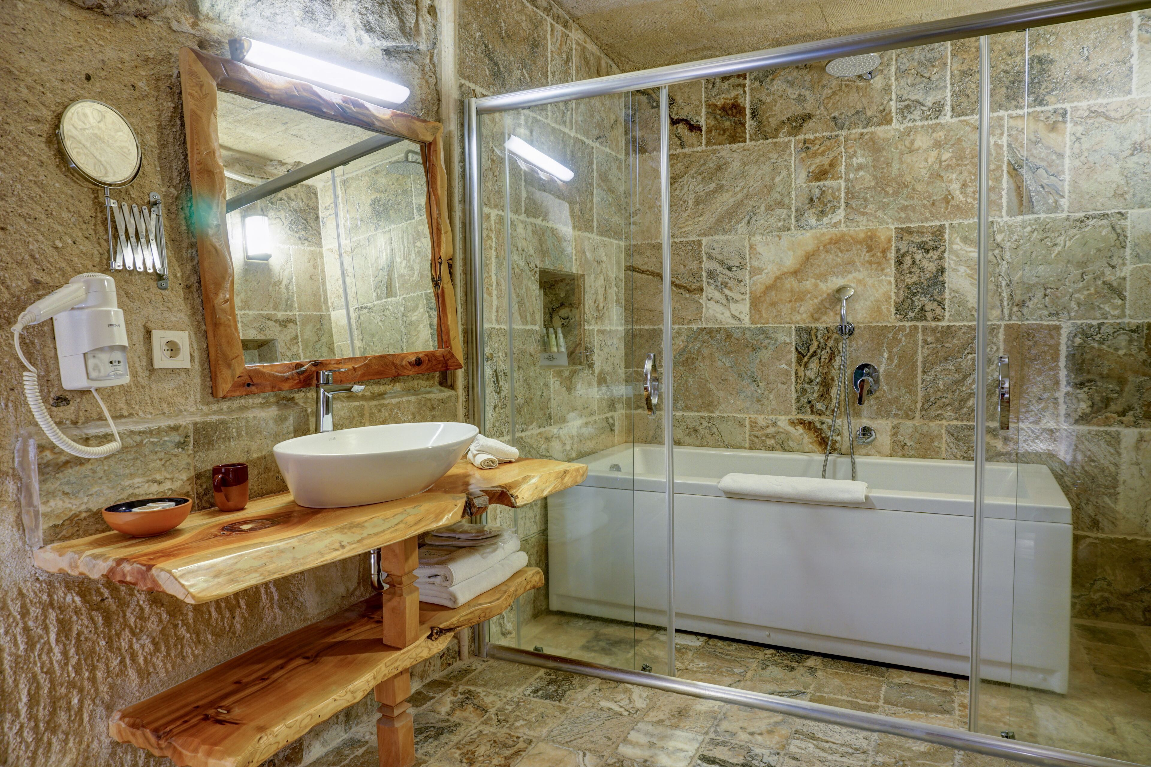 A bathroom with a stone wall, a wooden vanity, a large sink, a bathtub, and a glass-enclosed shower.