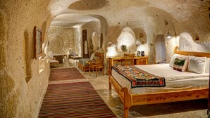 Um quarto rústico e acolhedor com cama de madeira, uma pequena escrivaninha e uma cadeira. O espaço apresenta um teto e paredes com aspecto de caverna, criando uma atmosfera natural e intimista.