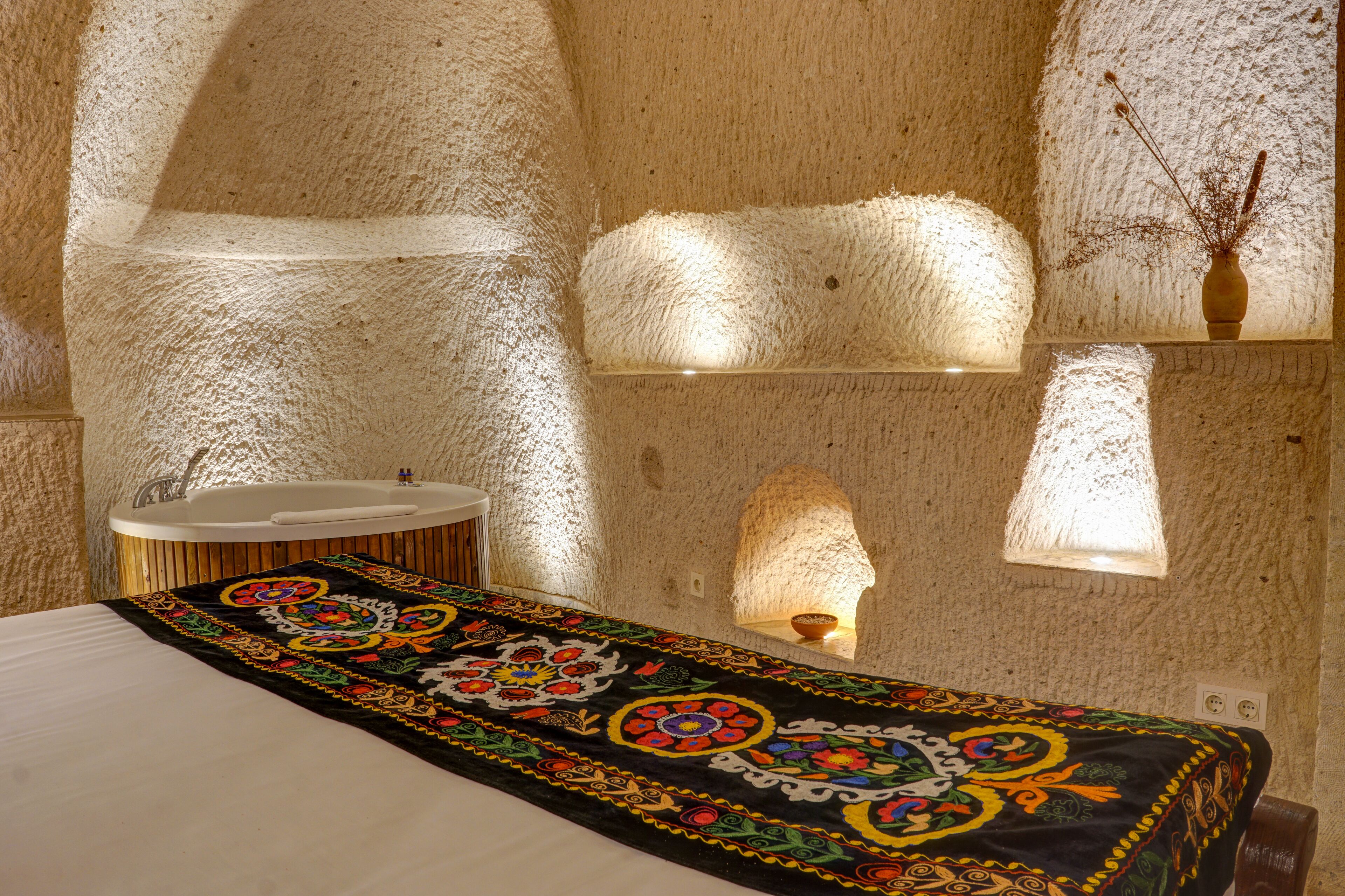 A room with a stone wall, a bed with a colorful bedspread, a sink, and a vase with branches.