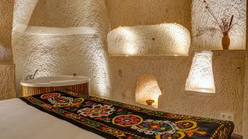 A room with a stone wall, a bed with a colorful bedspread, a sink, and a vase with branches.