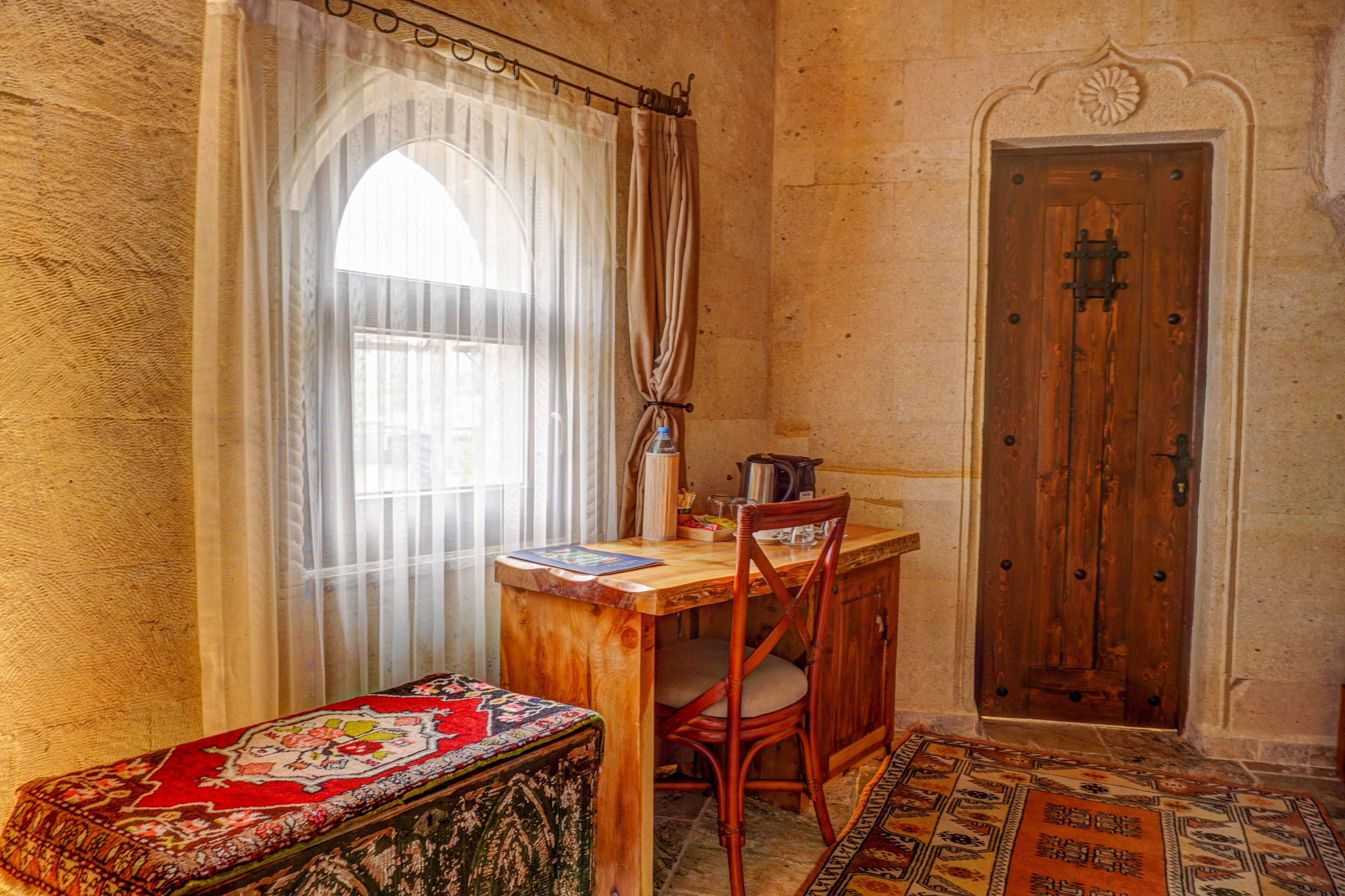 A room with a wooden desk, a chair, a patterned bench, a door, and a window with sheer curtains.
