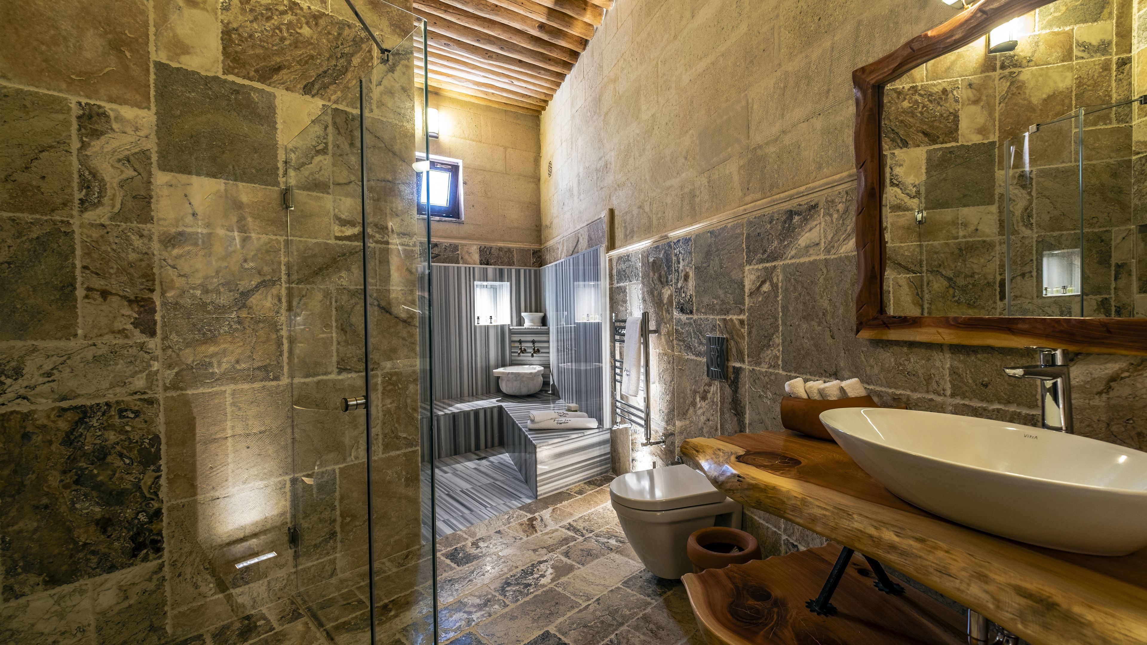 A bathroom with stone walls, a glass shower enclosure, a wooden vanity with a large sink, and a mirror.