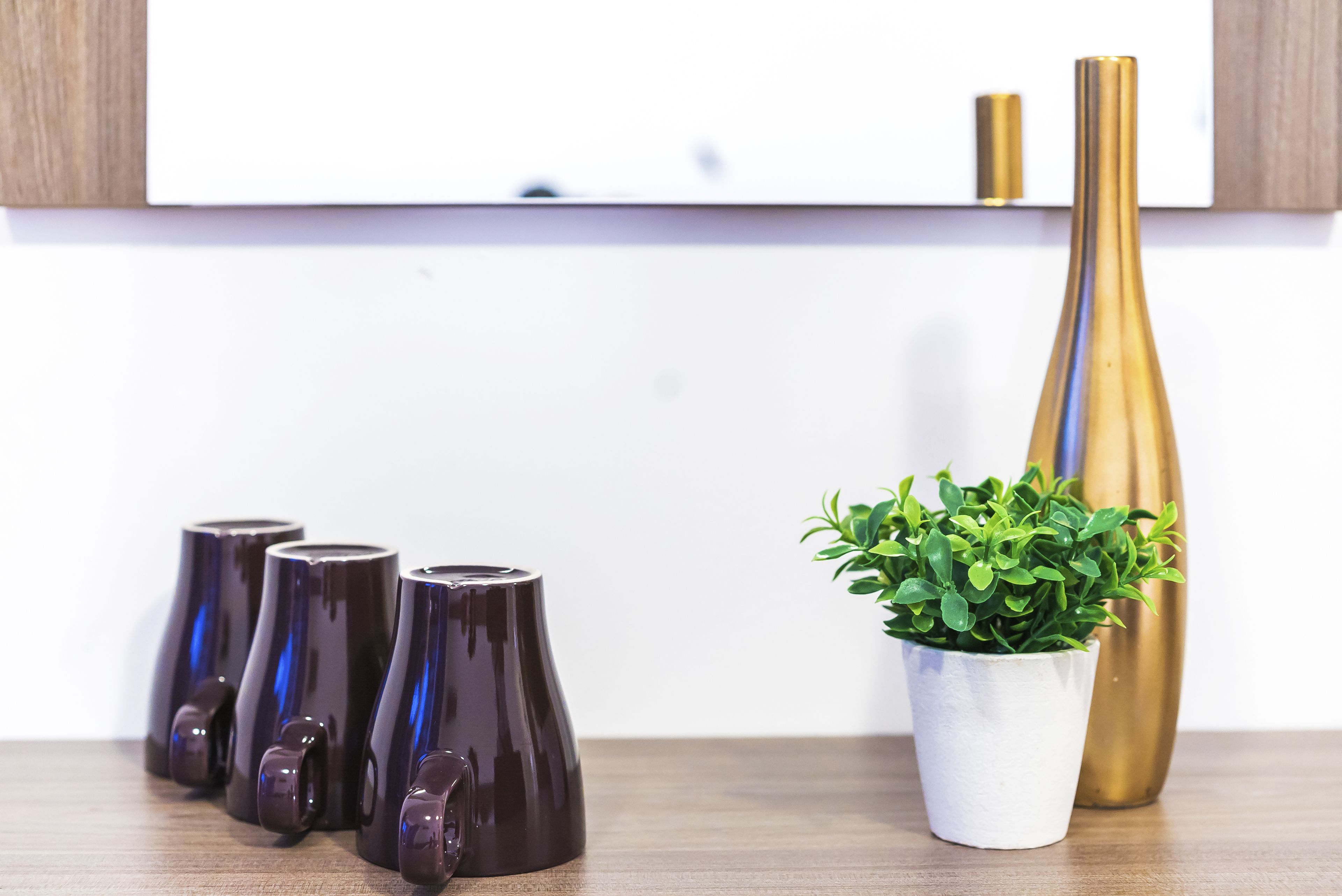 A row of dark vases on a wooden surface, a small potted plant, and two tall vases, one gold and one brown, against a white wall.