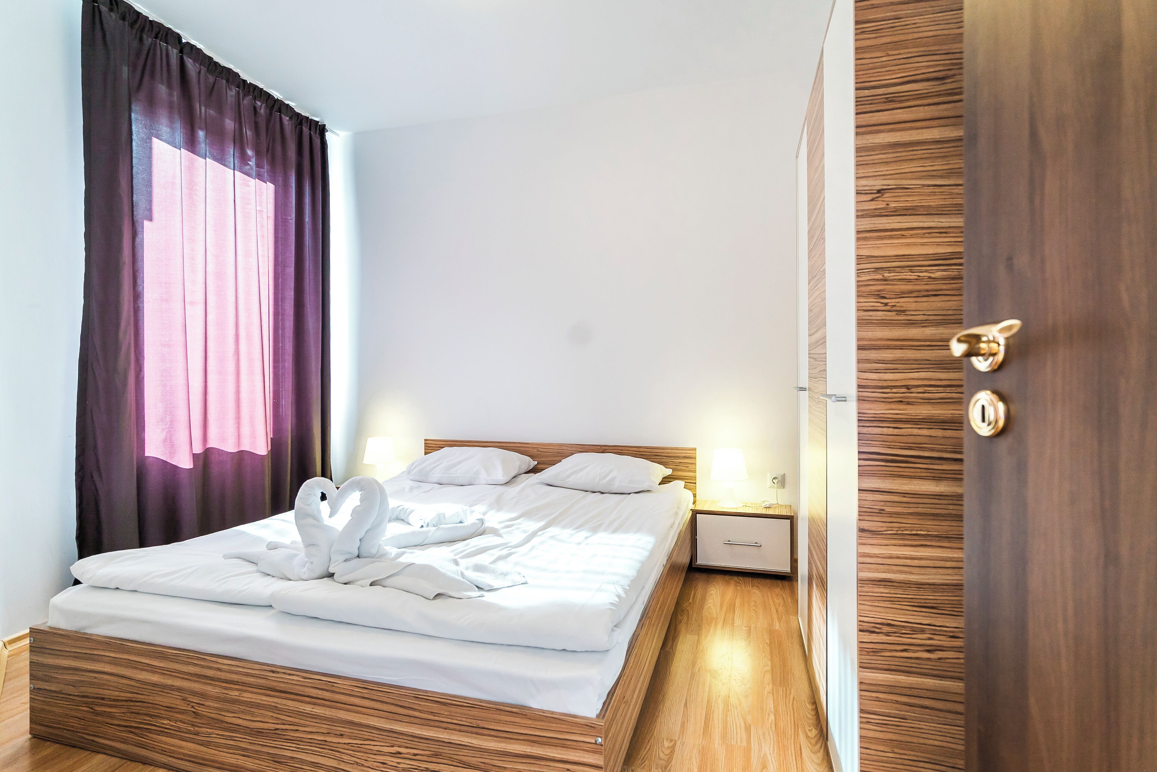 Una cama bien hecha con sábanas blancas y un arreglo de almohadas con forma de cisne. Se observa un cabecero de madera, una mesita de noche con lámpara, y una puerta de madera con pomo de latón.