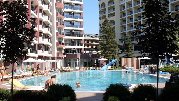 Ein Schwimmbecken mit Liegestühlen und Sonnenschirmen, umgeben von Grünflächen und hohen Gebäuden.