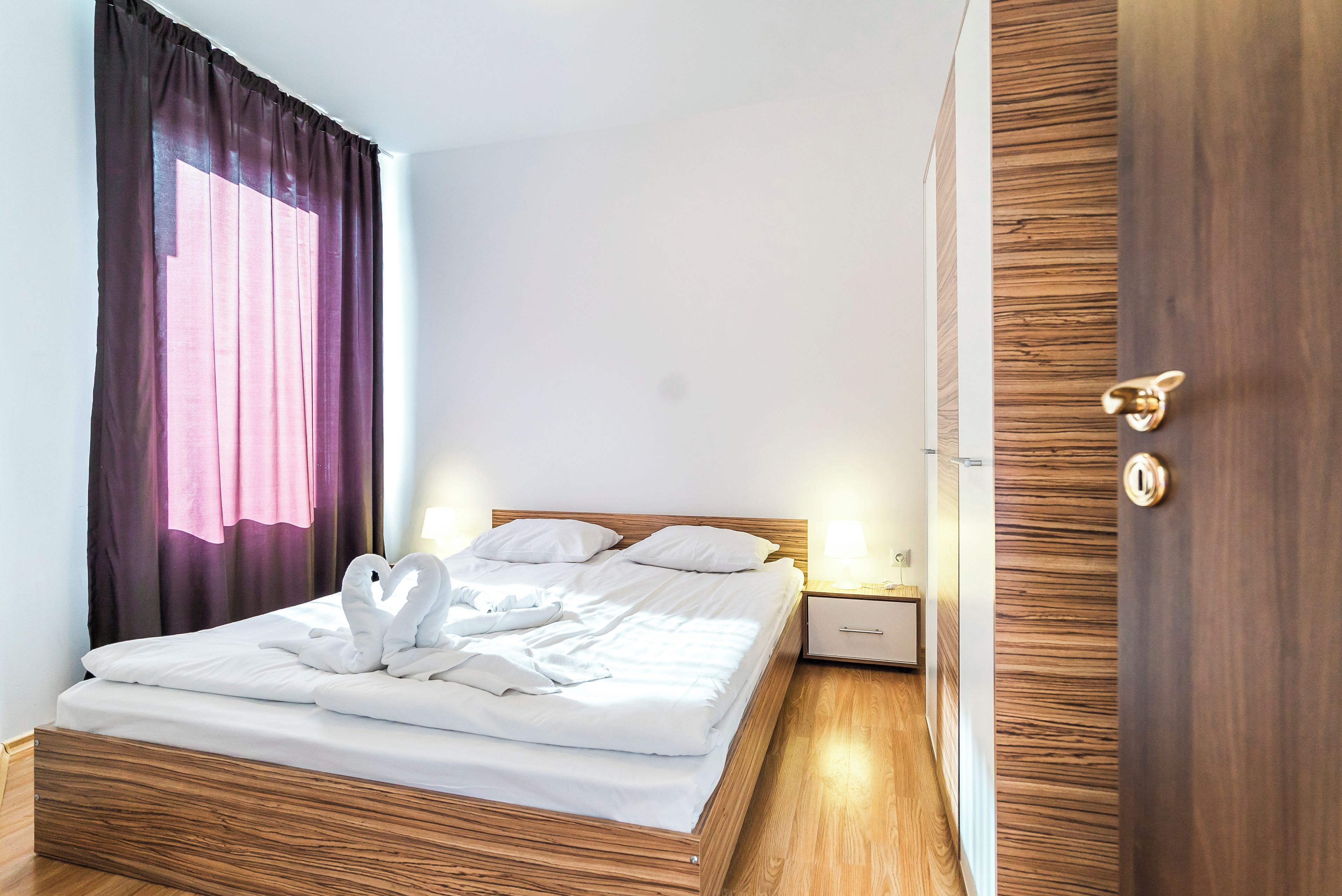 Un lit bien fait, avec du linge de lit blanc et un coussin décoratif en forme de cygne, une tête de lit en bois, une table de chevet avec une lampe et une porte en bois dotée d’une poignée en laiton.