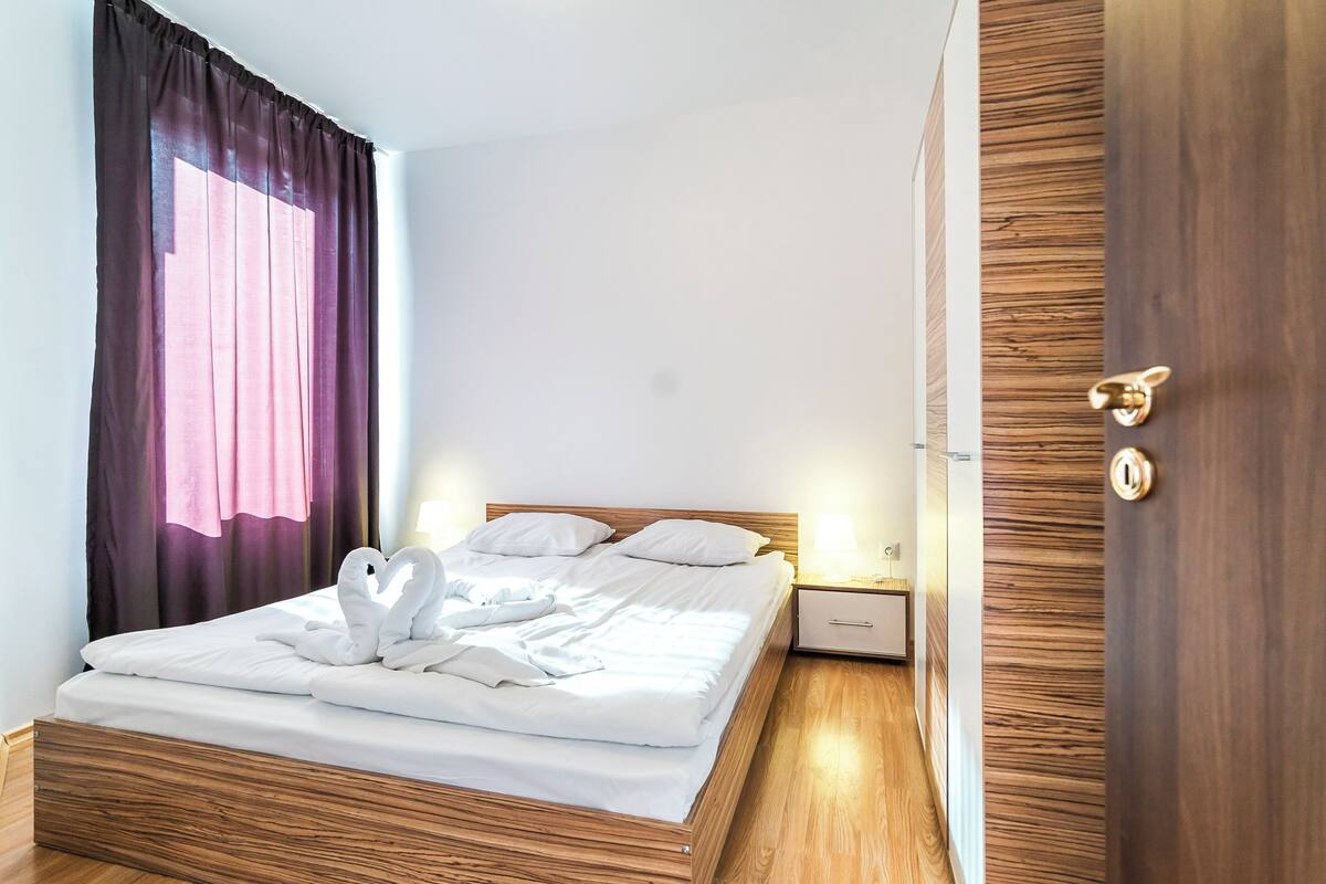 A neatly made bed with white linens and two white swan-shaped pillows, a wooden headboard, a nightstand with a lamp, and a wooden door with a brass handle.