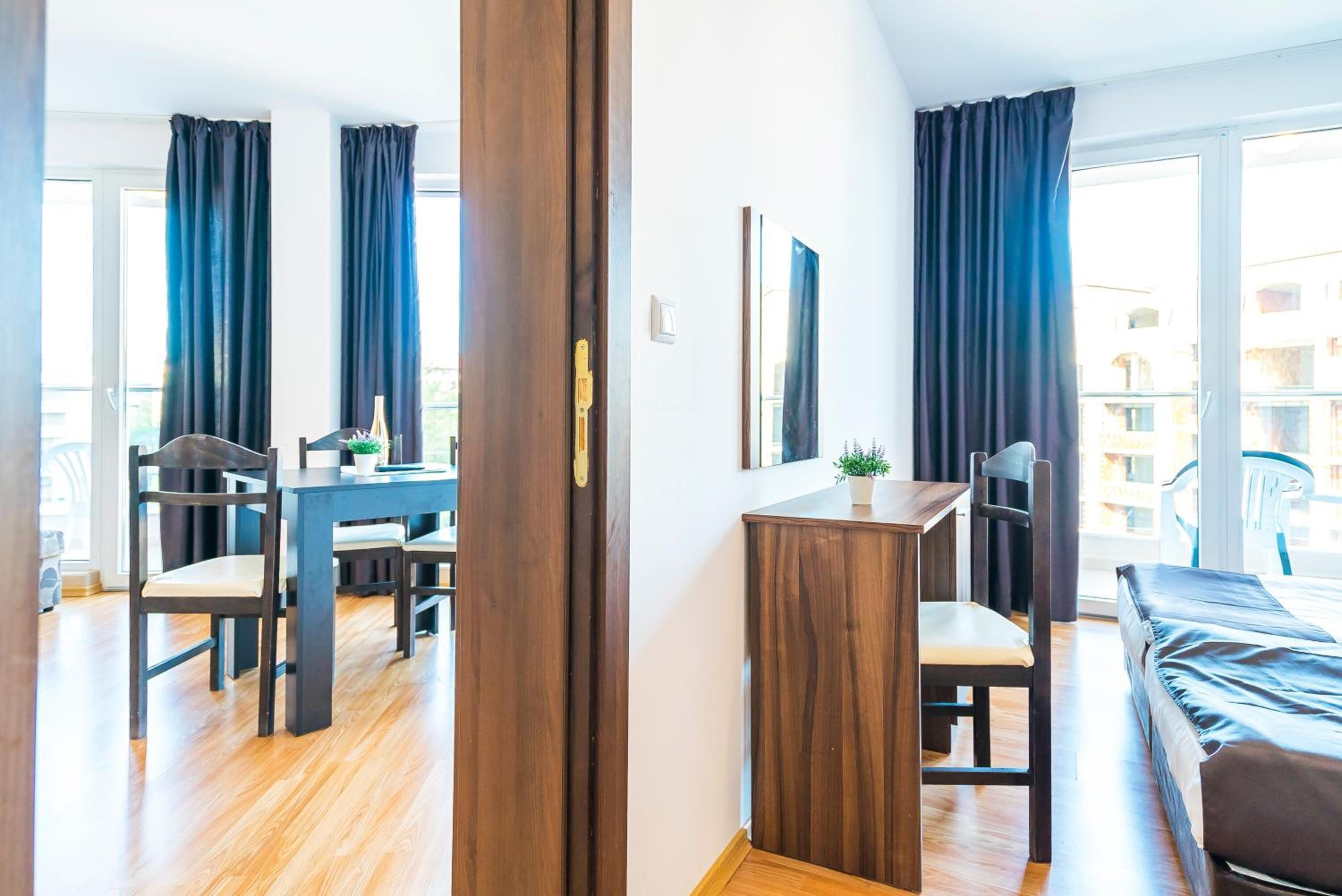 A modern room with a wooden floor, a dining table with chairs, a small cabinet, a bed, and large windows with curtains.