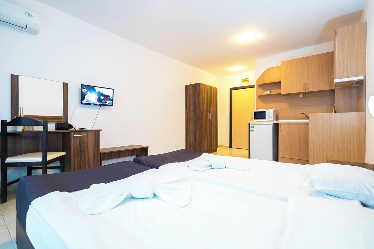 A hotel room with a bed, a desk, a chair, a TV, and a kitchenette.