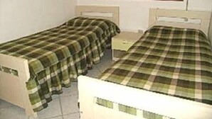 Une petite chambre avec deux lits simples, chacun recouvert d’une couverture à carreaux verts et blancs. Il y a une petite table de chevet entre les lits et une fenêtre agrémentée d’un rideau décoratif.