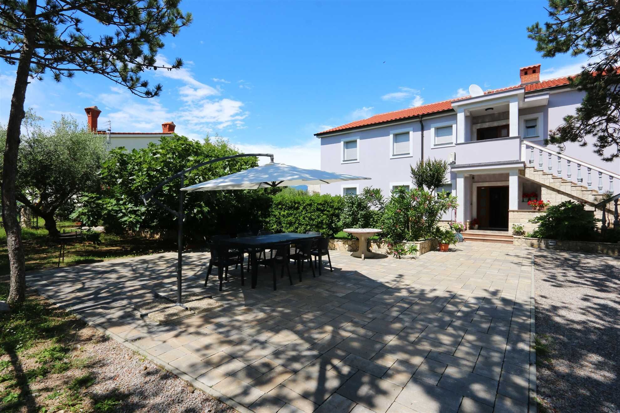 A courtyard with a patio table, chairs, and an umbrella. There is a building with a red roof and a staircase leading to a balcony.