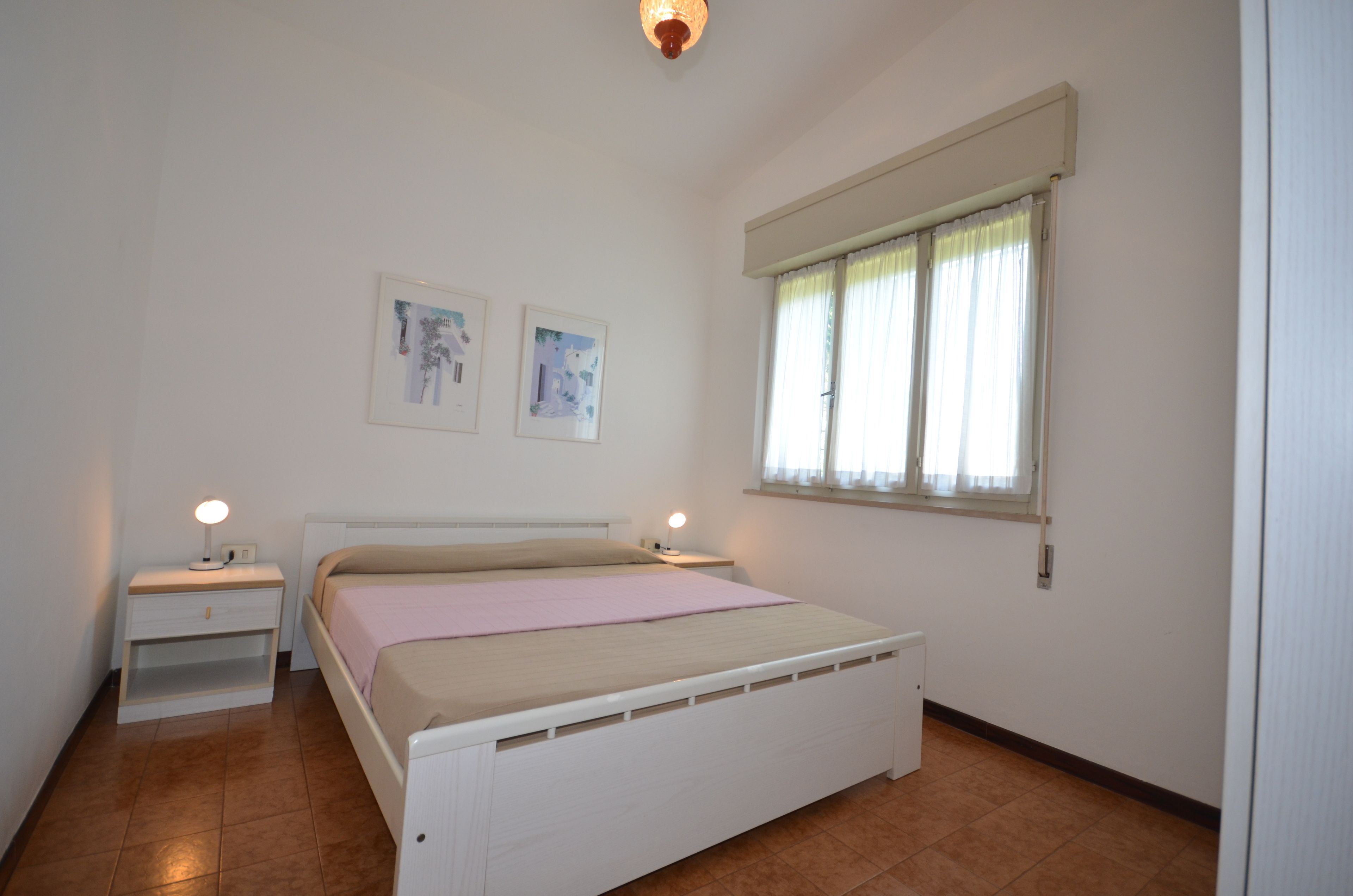 A bedroom with a single bed, two bedside tables with lamps, a window with curtains, and two wall-mounted pictures.