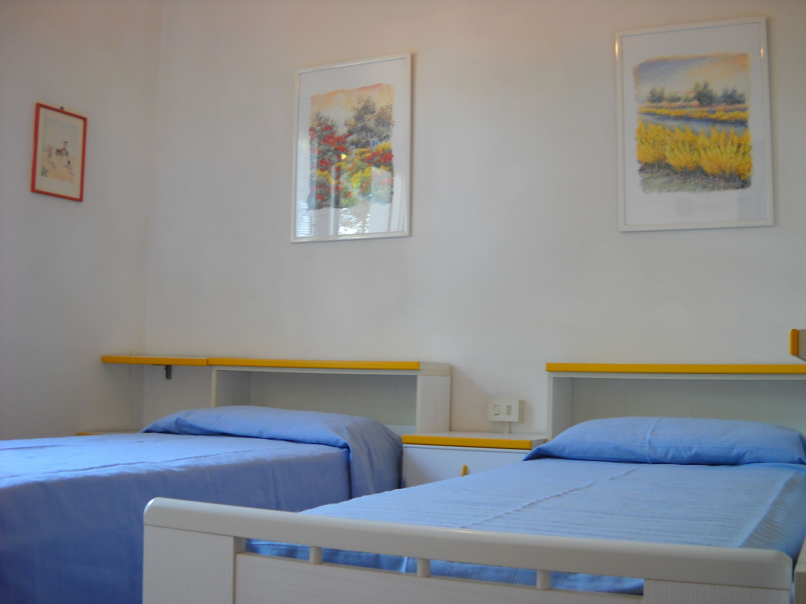 Una habitación con dos camas individuales, cada una con colchón y almohada azules, y cabeceros blancos. Hay dos obras de arte enmarcadas en la pared, una que representa una escena abstracta colorida y la otra un paisaje con follaje amarillo.