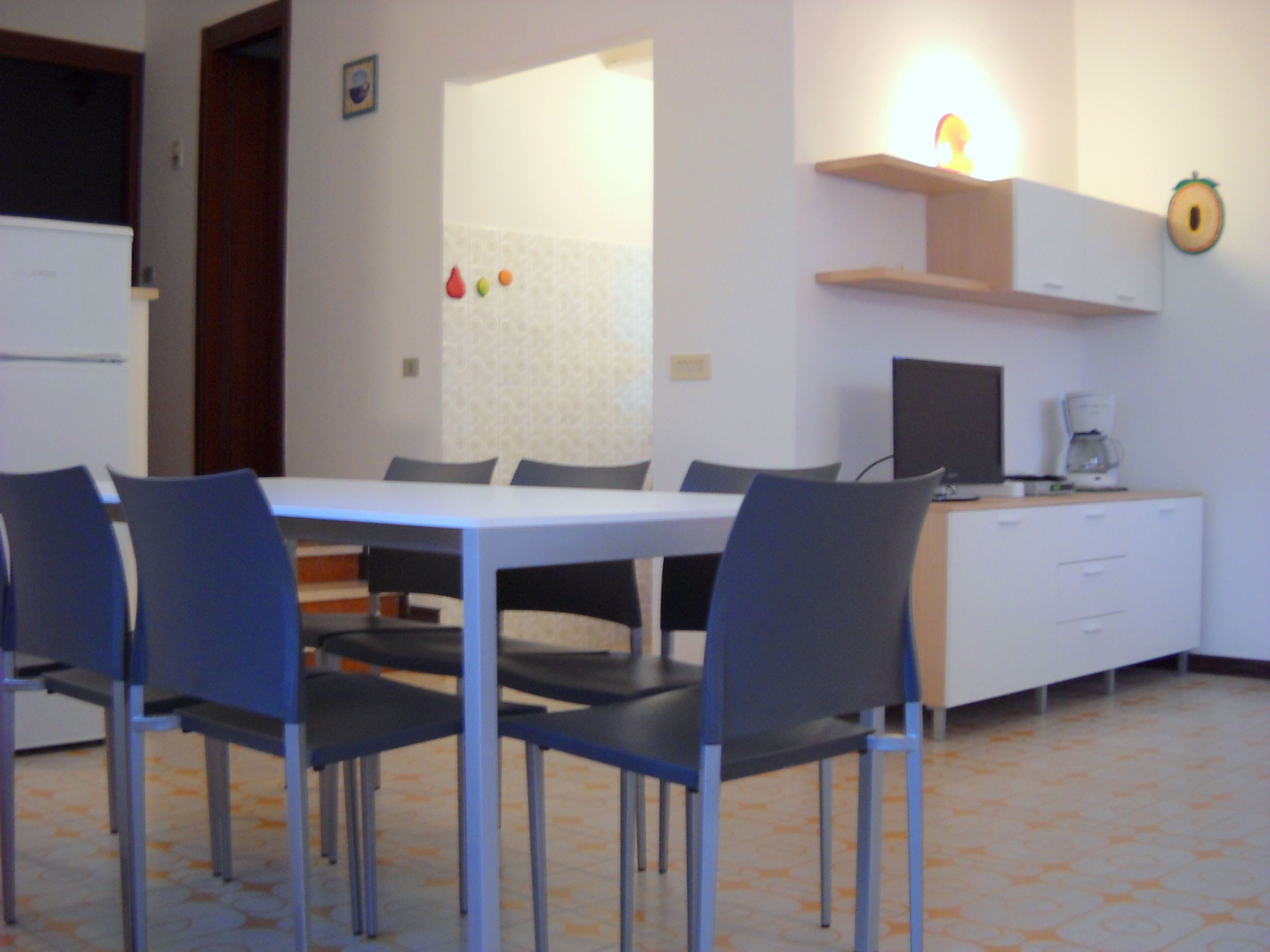 A dining area with a white table and blue chairs, a small kitchen with a coffee maker, and a wall clock.