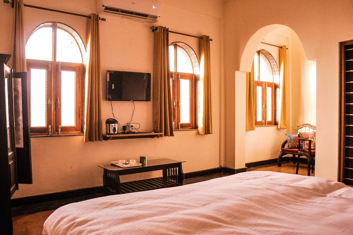 A hotel room with a large bed, a small table with a tray, and two windows with curtains.