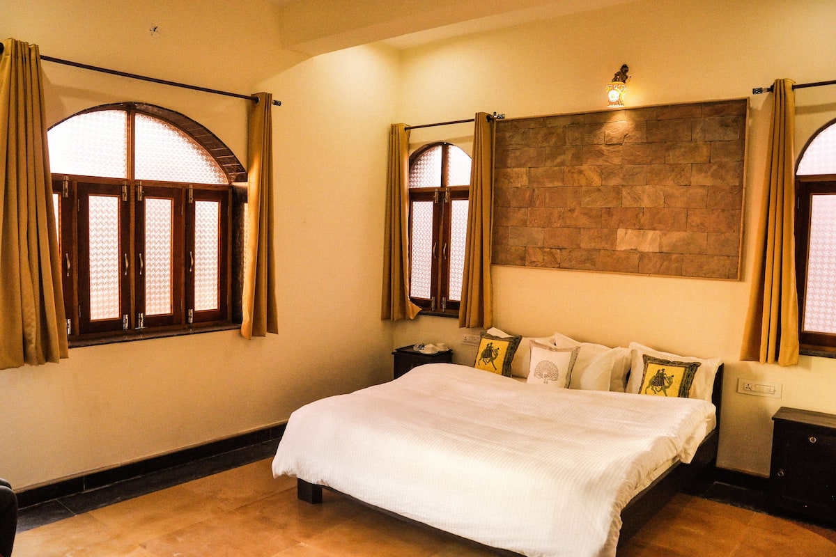 A bedroom with a large bed, two windows with curtains, a stone wall, and a nightstand with a lamp.