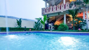 A swimming pool with a fountain, surrounded by greenery and a building with multiple balconies. - Hotel Vamdev Fort (Pushkar)
