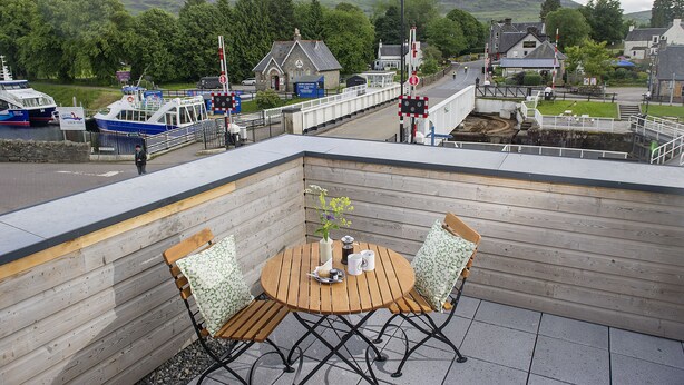 A wooden deck with a round table and chairs, overlooking a harbor with boats and a railway crossing.