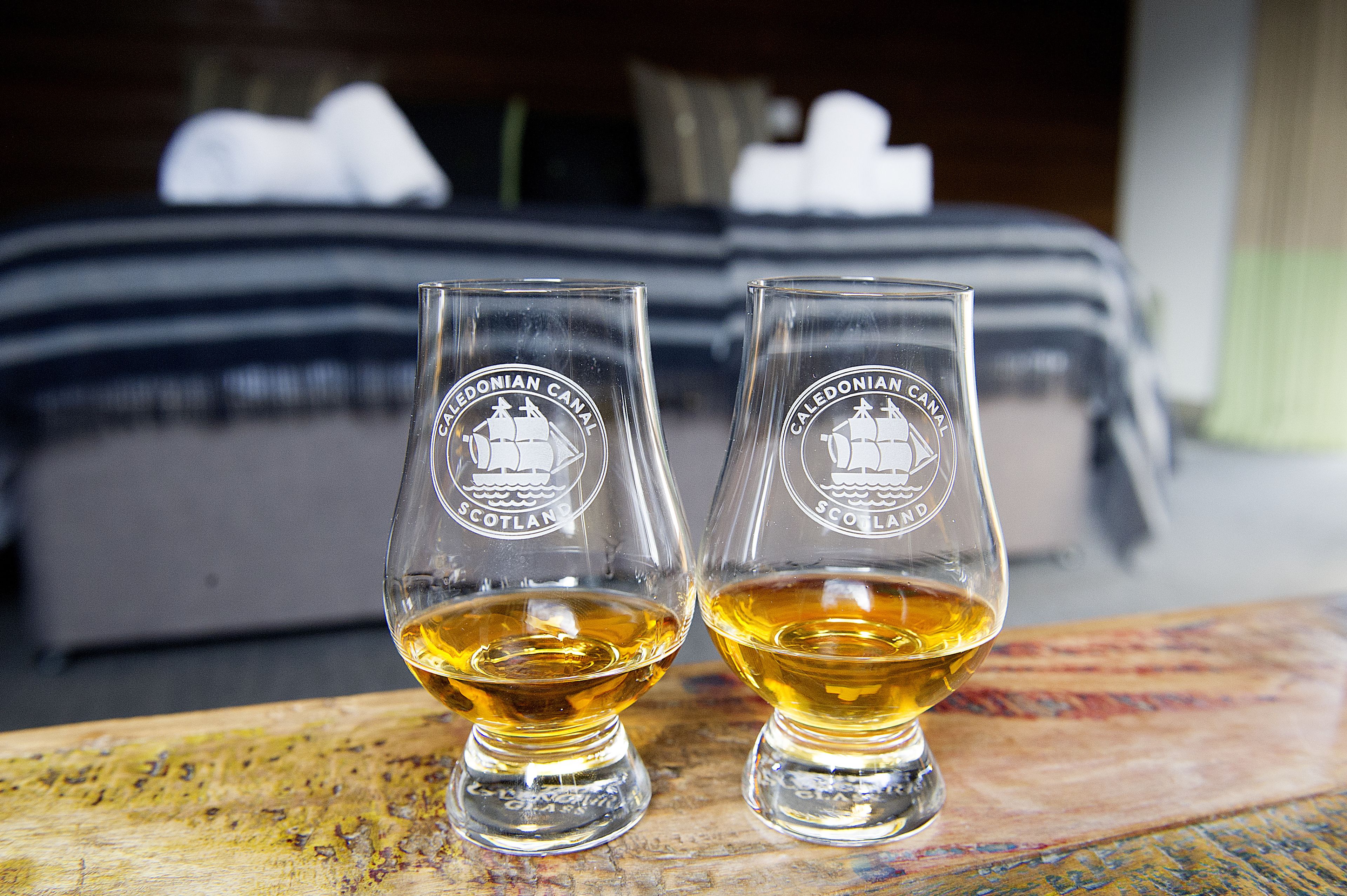 Two glasses of Caledonian Canal whisky on a wooden table with a bed in the background.